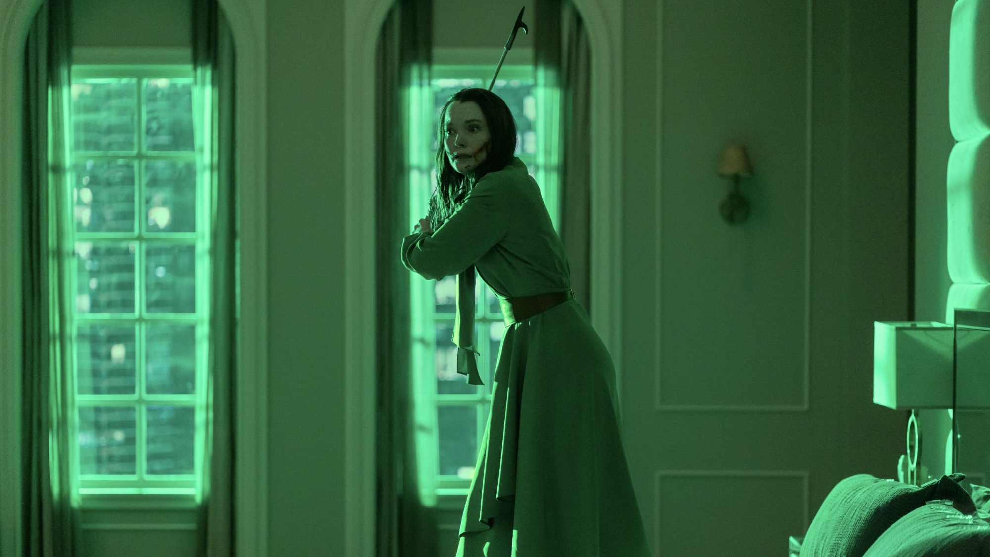 Une femme vêtue d’une robe verte se tient sur un lit dans une pièce éclairée en vert, tenant un tisonnier comme une arme.