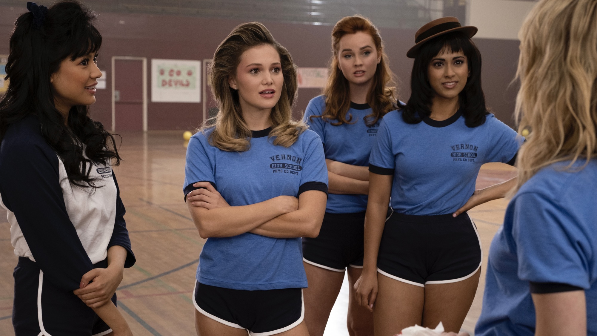 Un groupe de jeunes femmes en tenue de sport et coiffées des années 80 dans le gymnase d'un lycée.