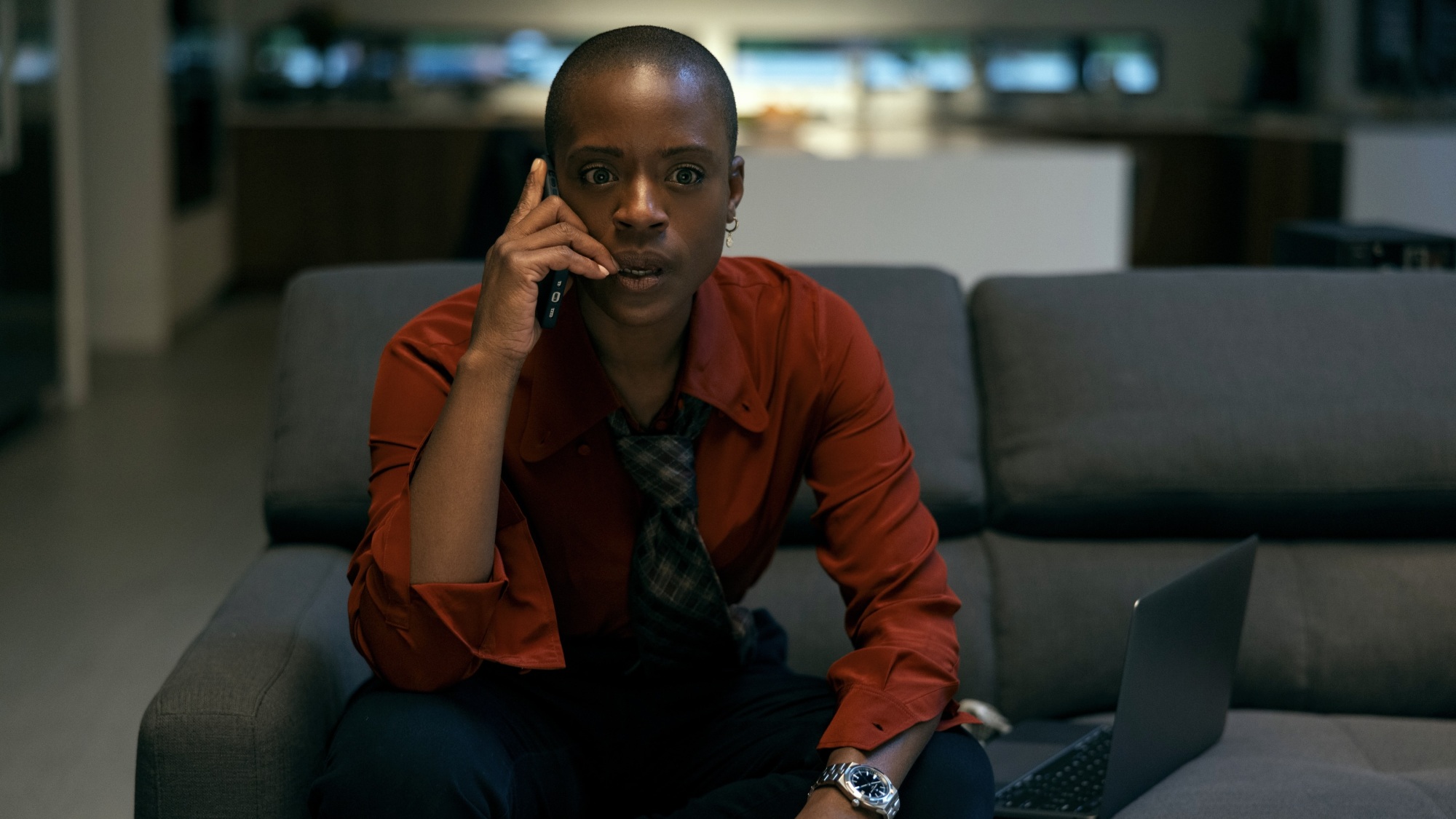 Une femme vêtue d'une chemise boutonnée rouge est assise sur un canapé gris tout en parlant au téléphone.