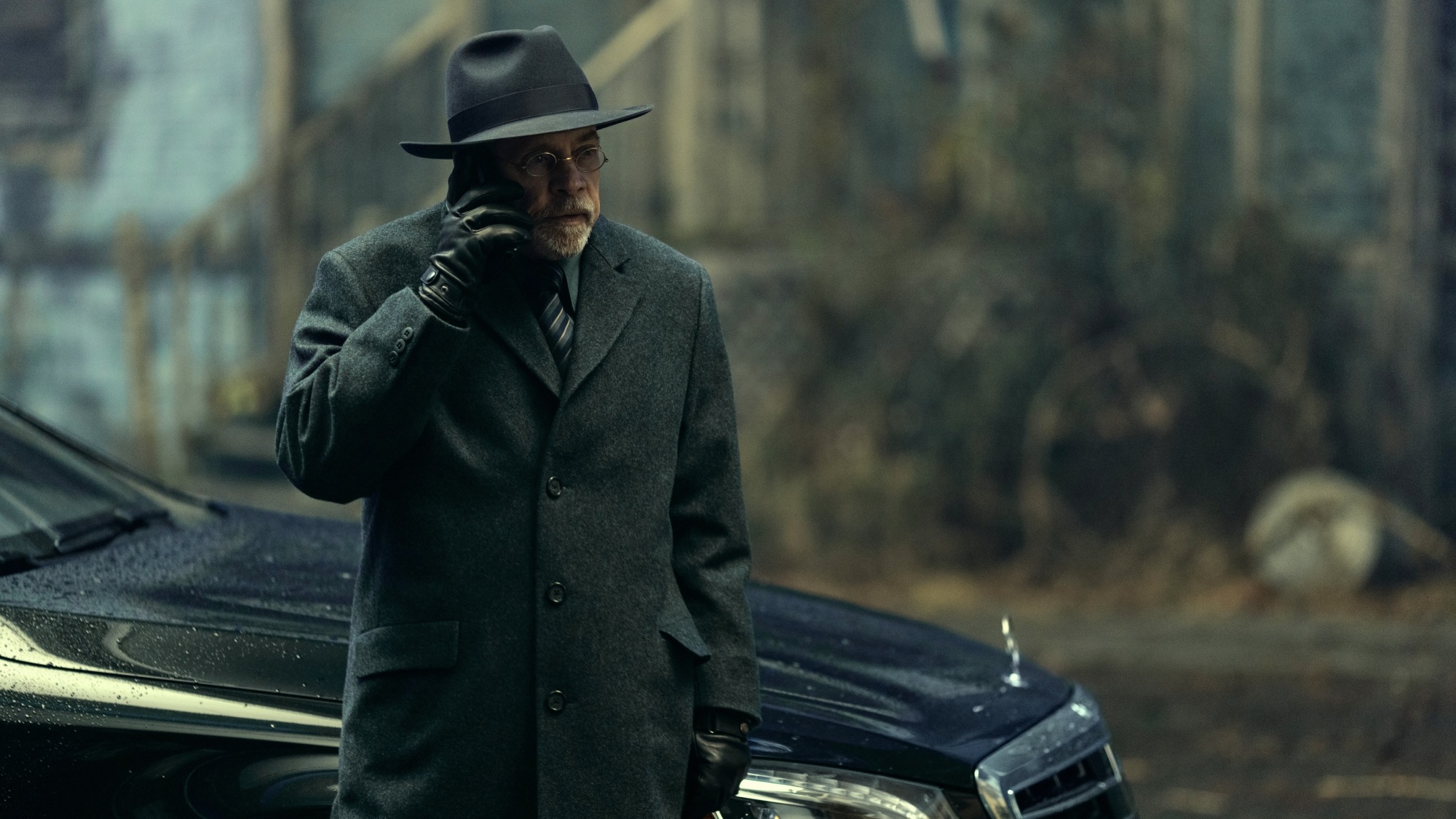 Un homme vêtu d'un manteau sombre, d'un chapeau et de gants parle au téléphone alors qu'il se tient à côté d'une voiture noire.