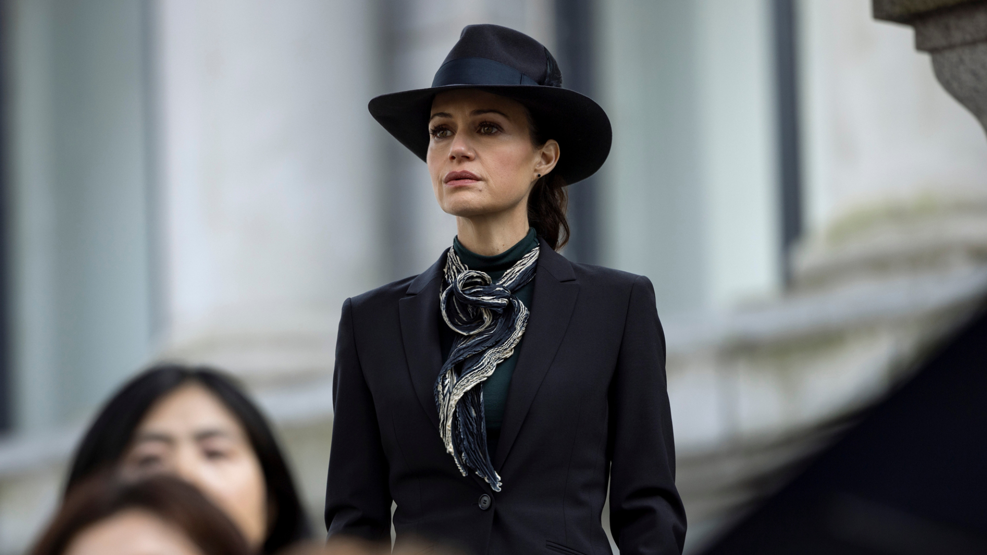 Une femme en costume noir et chapeau se tient parmi une foule devant un bâtiment public.