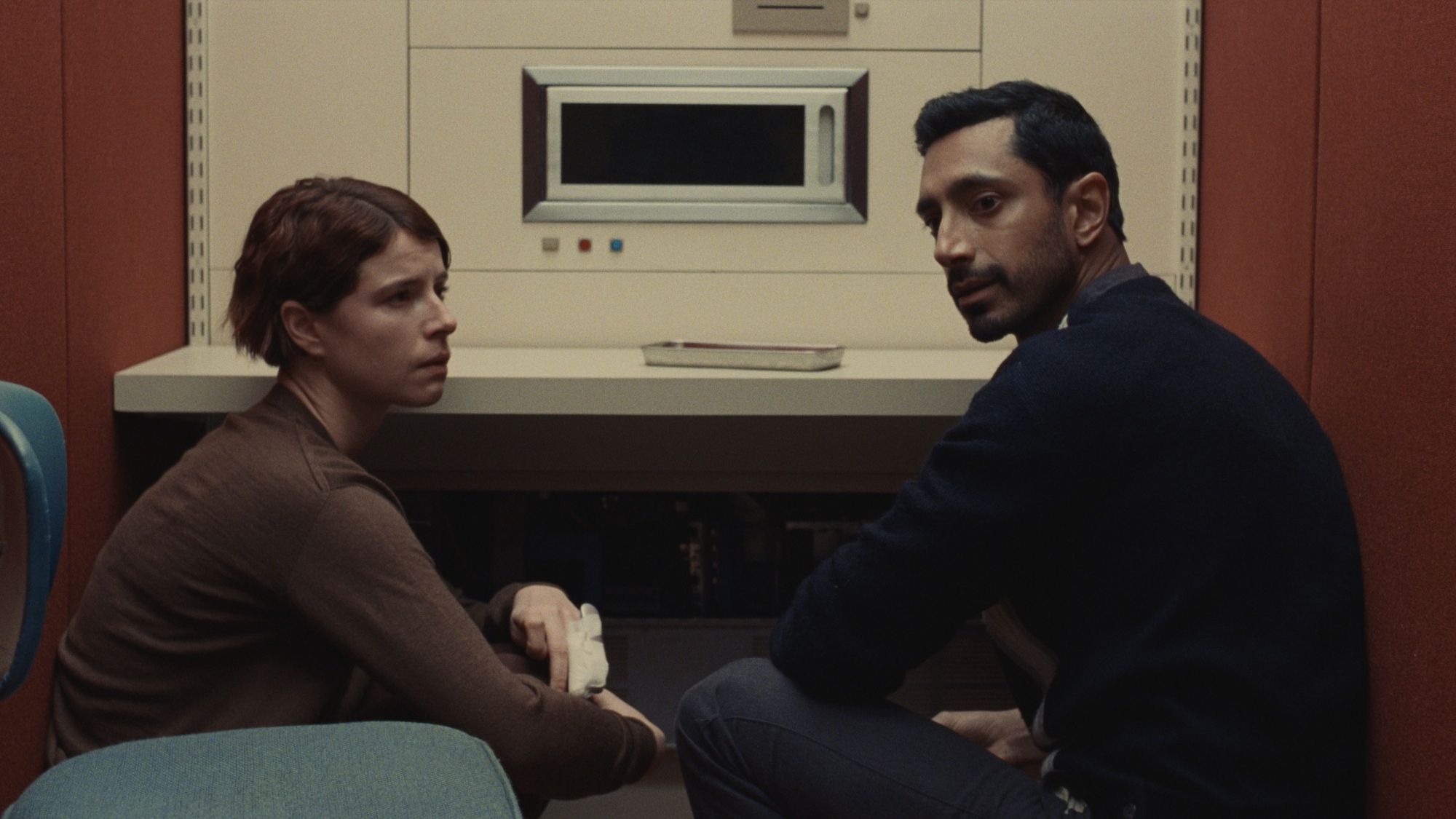 Un homme et une femme dans une pièce rouge, regardant une machine futuriste qui ressemble à un micro-ondes.
