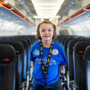 Un enfant debout dans l’allée centrale d’un avion, portant une chemise bleue Make-A-Wish. 