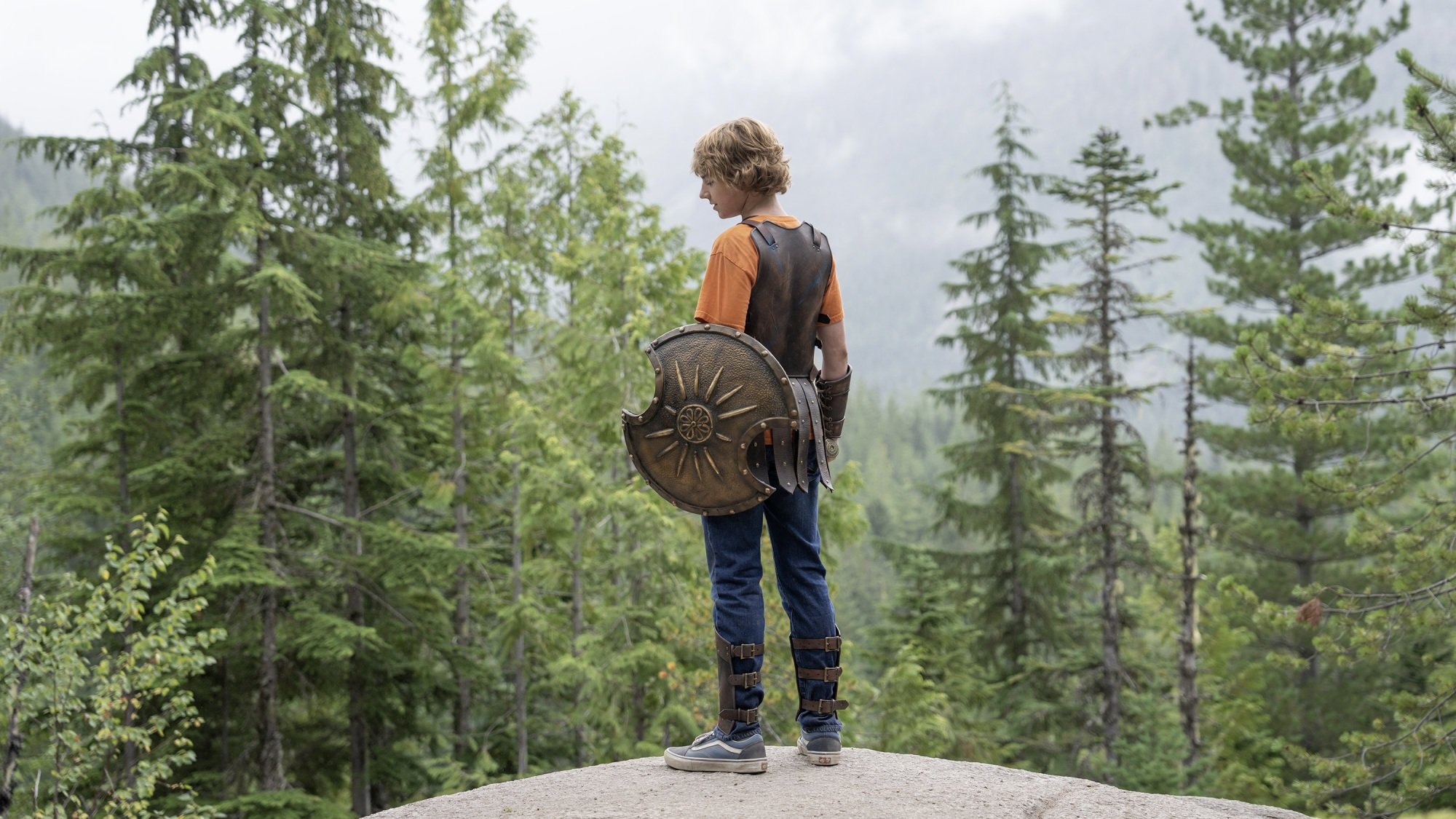 Un jeune garçon en armure grecque et un T-shirt orange se tient au sommet d’une colline surplombant une forêt.