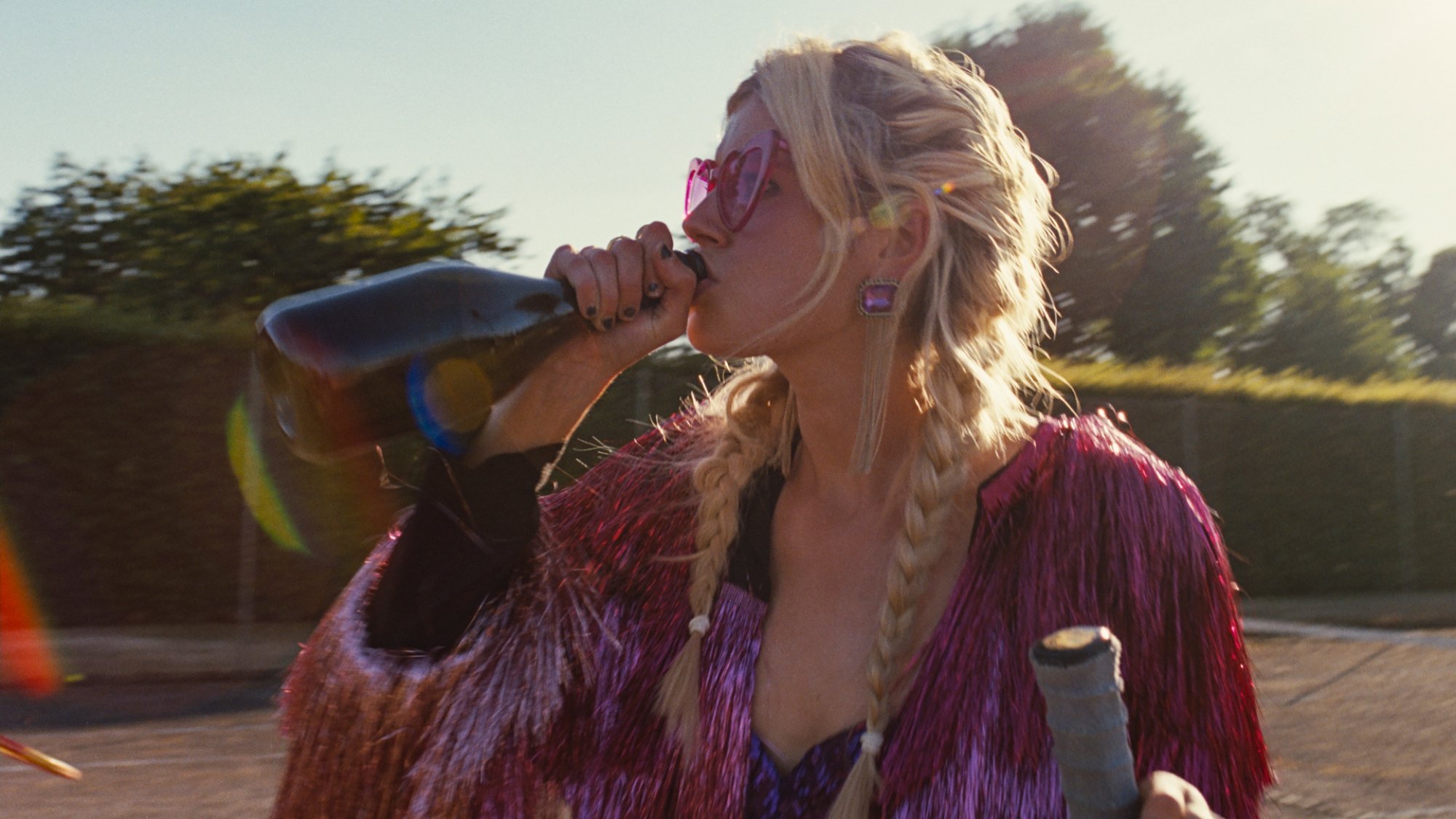 Une jeune femme portant des lunettes de soleil rose vif et une veste en guirlandes boit du champagne sur un court de tennis.