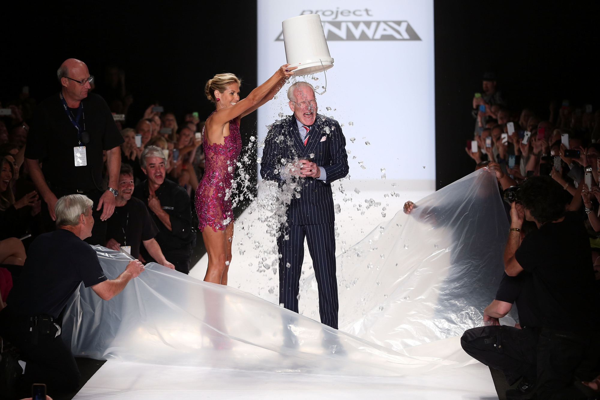 Heidi Klum (dans une robe à paillettes rose foncé) verse un seau d'eau glacée sur Tim Gunn (en costume) lors du spectacle final de la saison 13 de Project Runway le 5 septembre 2014. Ils sont sur une piste blanche.