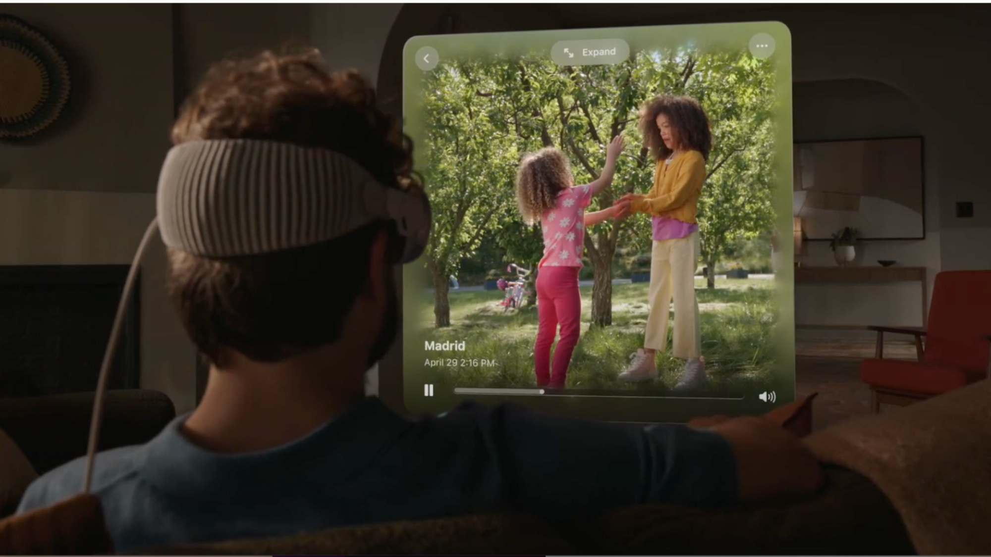 Un homme blanc aux cheveux bouclés portant l’Apple Vision Pro est vu de dos.  Il regarde une vidéo de deux filles (vraisemblablement ses filles) sur un écran projeté devant lui.