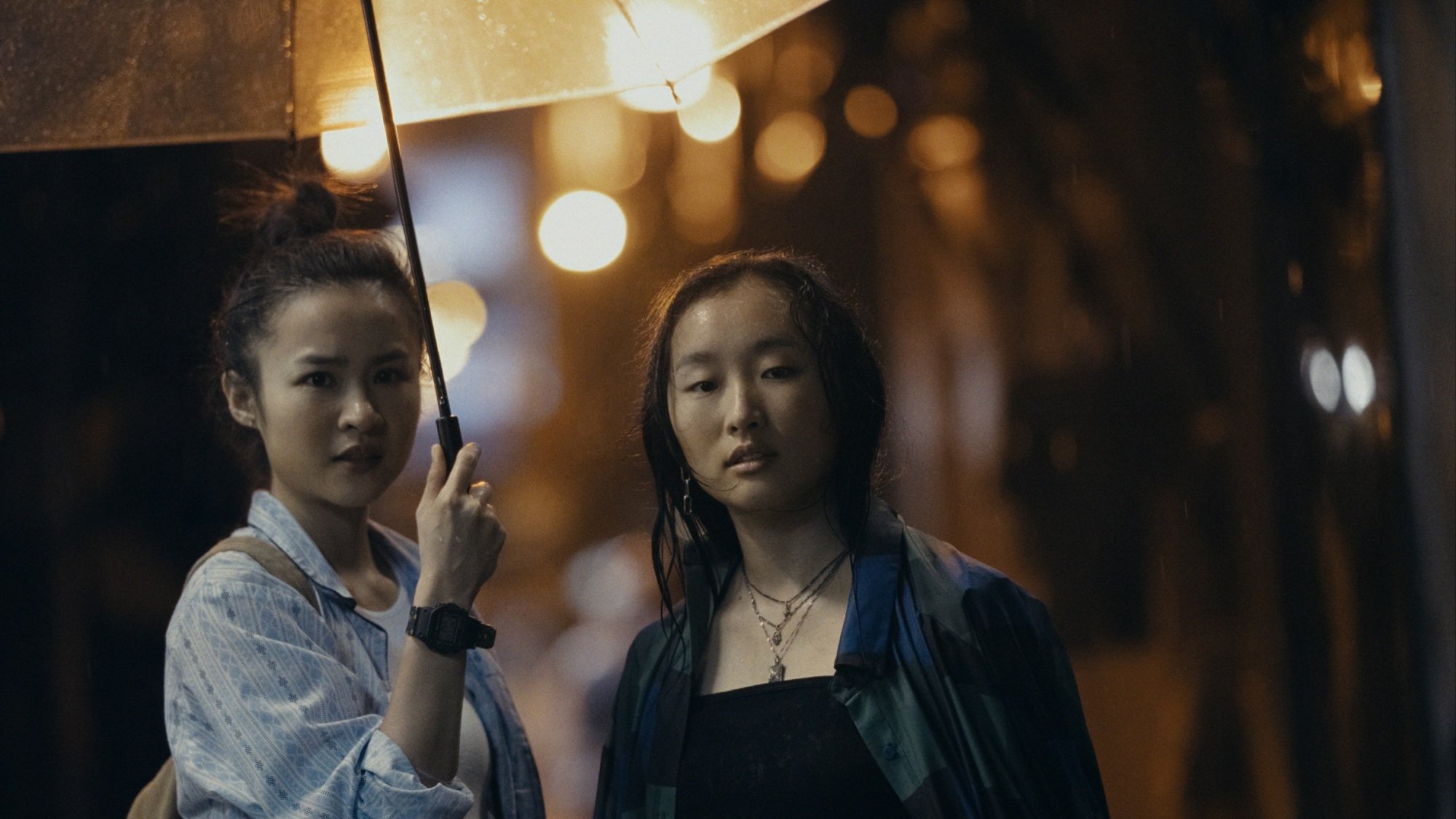 Deux femmes debout sous un parapluie transparent au milieu d’une rue pluvieuse.