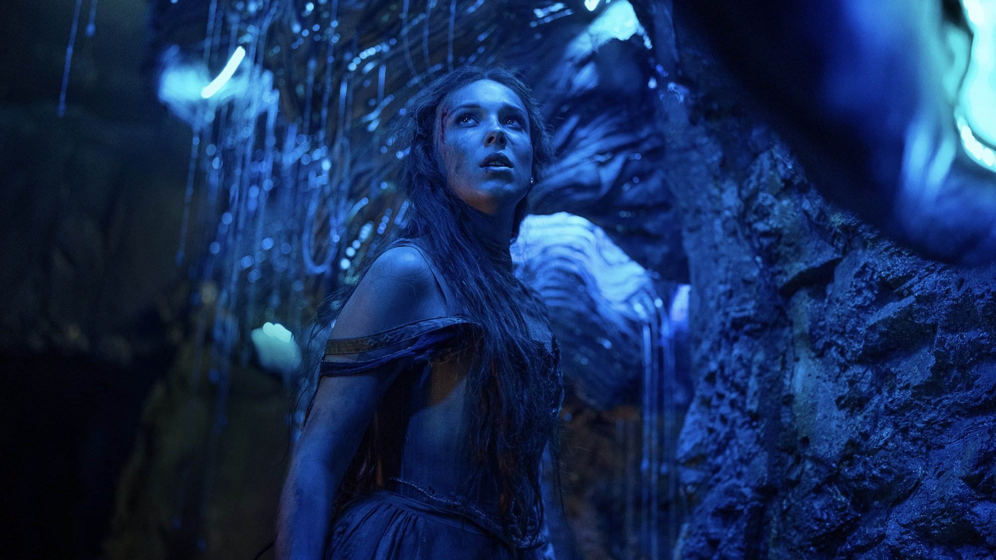 Une jeune femme se tient dans une grotte éclairée en bleu par des vers luisants.