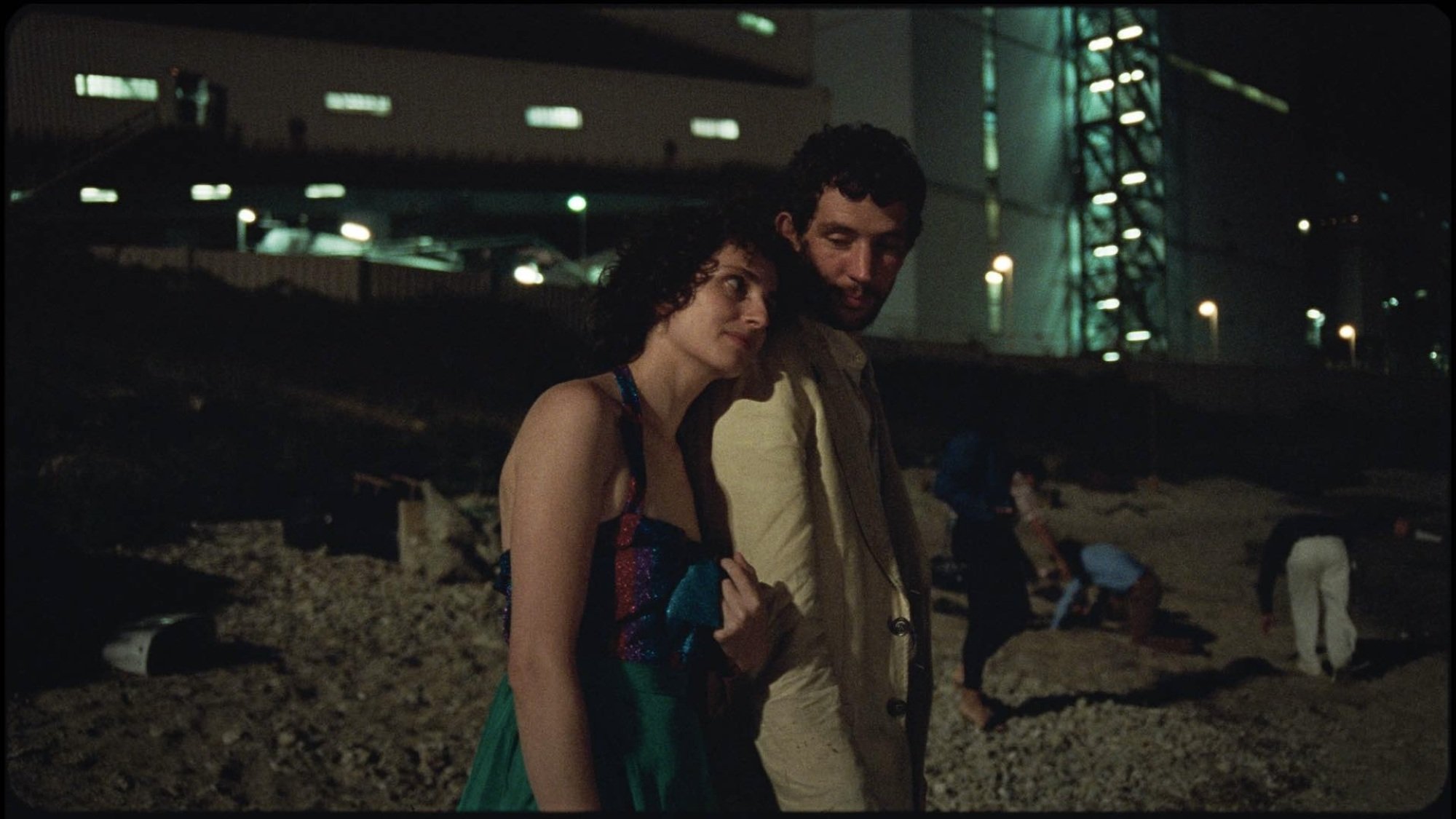 Une femme pose sa tête sur l'épaule d'un homme alors qu'ils se tiennent sur une plage en contrebas d'une usine.