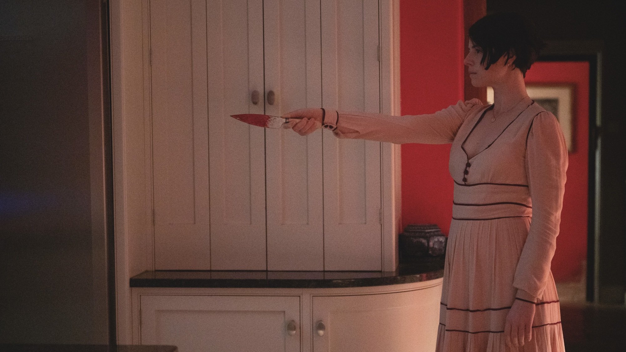 Une femme vêtue d’une robe rose pointe un couteau ensanglanté sur un intrus inconnu.
