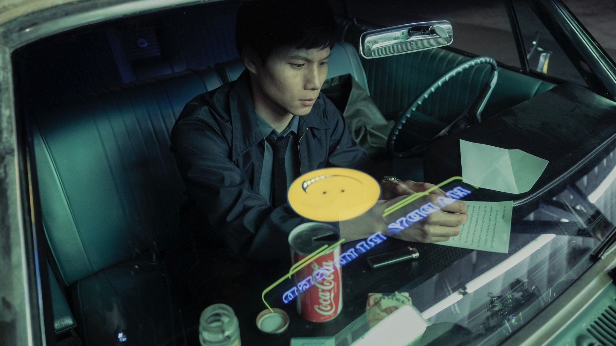 Un homme écrivant une lettre sur le siège avant d’une voiture ;  la lumière d'un panneau souriant jaune se reflète dans la fenêtre. 