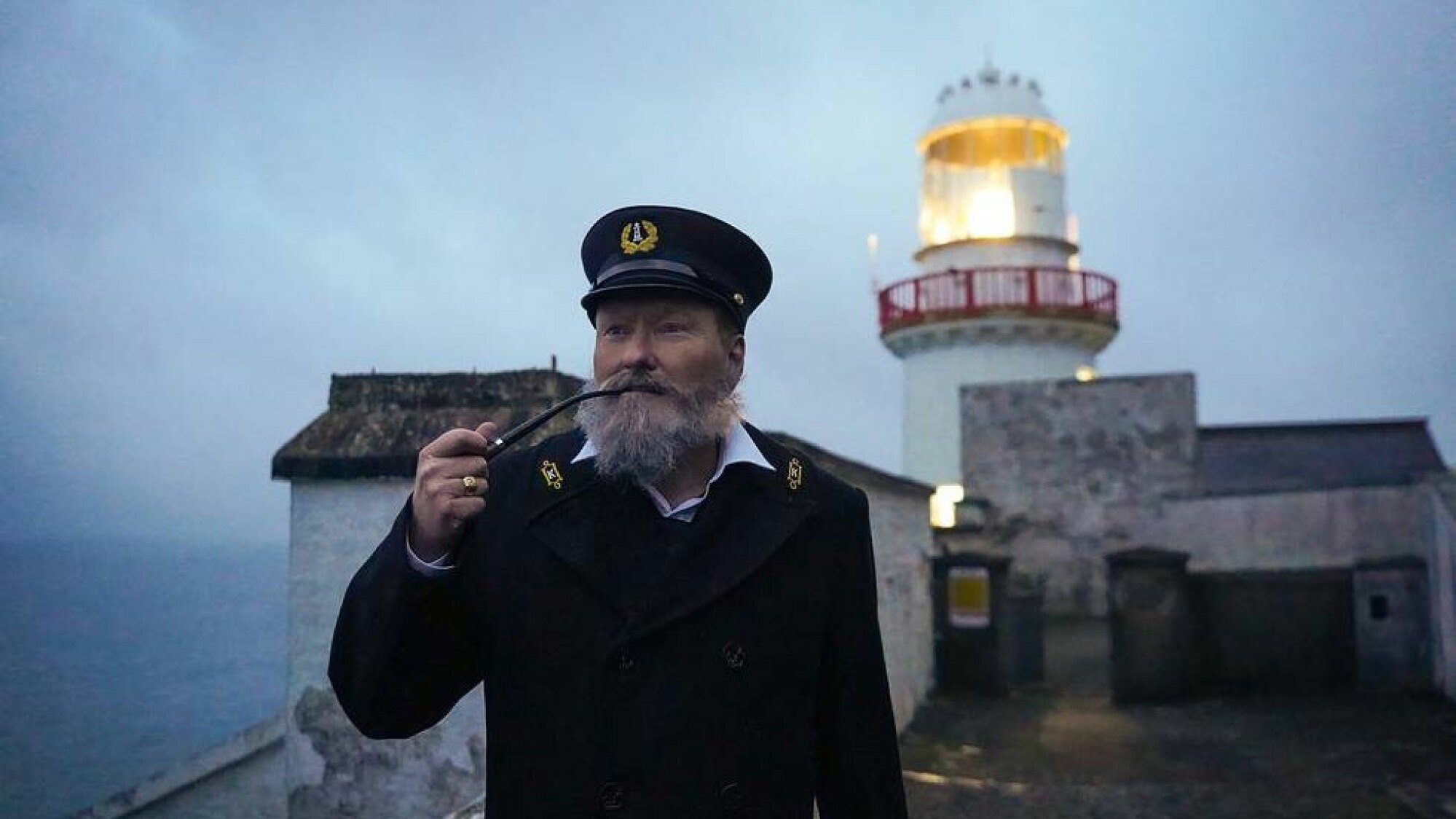 Un homme vêtu d’une vieille tenue de gardien de phare se tient devant un phare.
