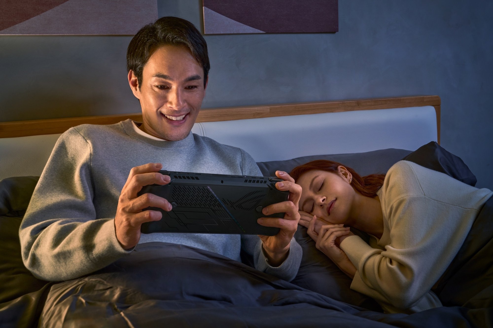 Homme au lit tenant un Asus ROG Ally X pendant qu'une femme dort à côté de lui