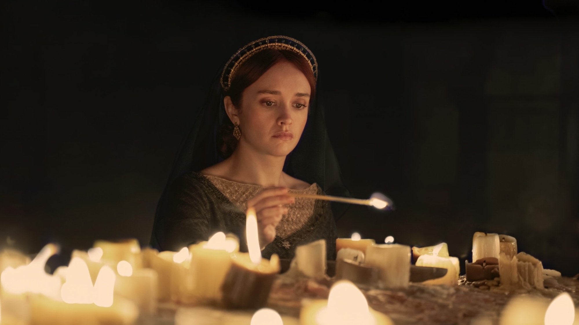 Alicent de "Maison du Dragon" allume des bougies sur un autel, vêtue d'une robe verte et d'une coiffe voilée noire.