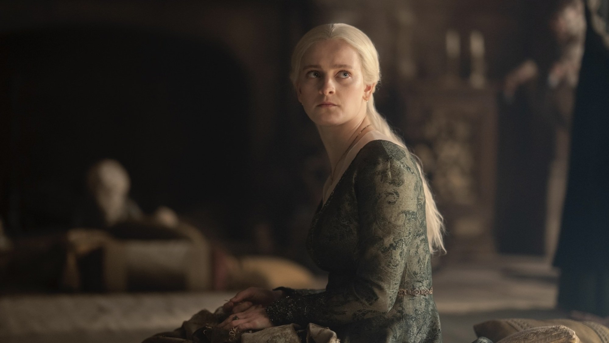 Helaena Targaryen est assise par terre dans une robe verte, levant les yeux de sa broderie.