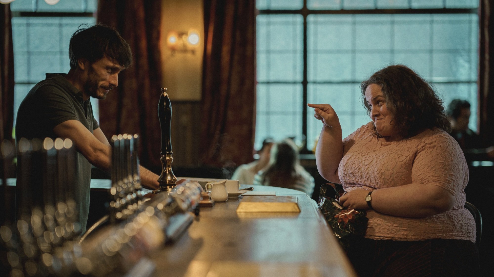 Une femme en chemise rose est assise dans un bar, désignant l'homme souriant qui s'occupe du bar.