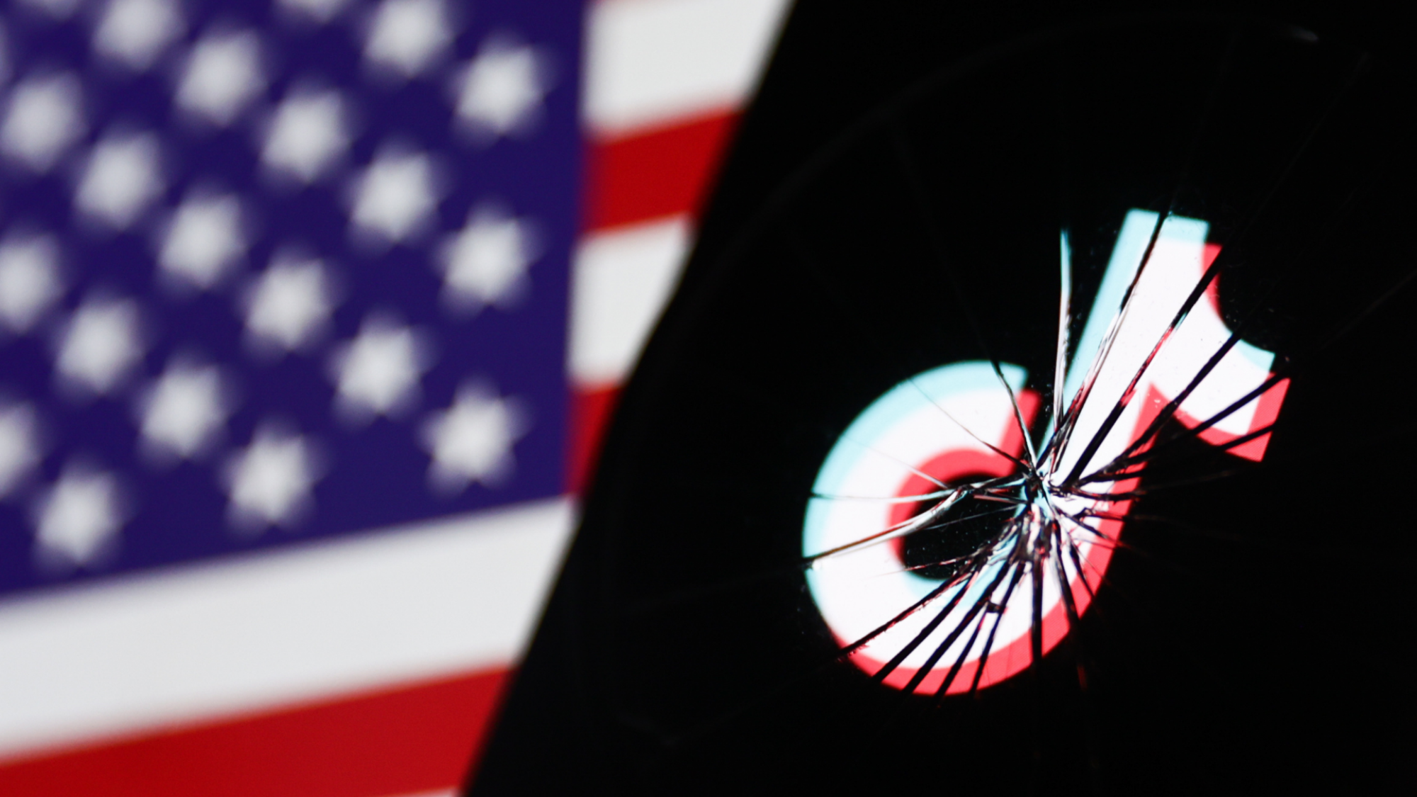 Le logo TikTok sur un écran de téléphone portable brisé, derrière lequel est visible le drapeau américain.