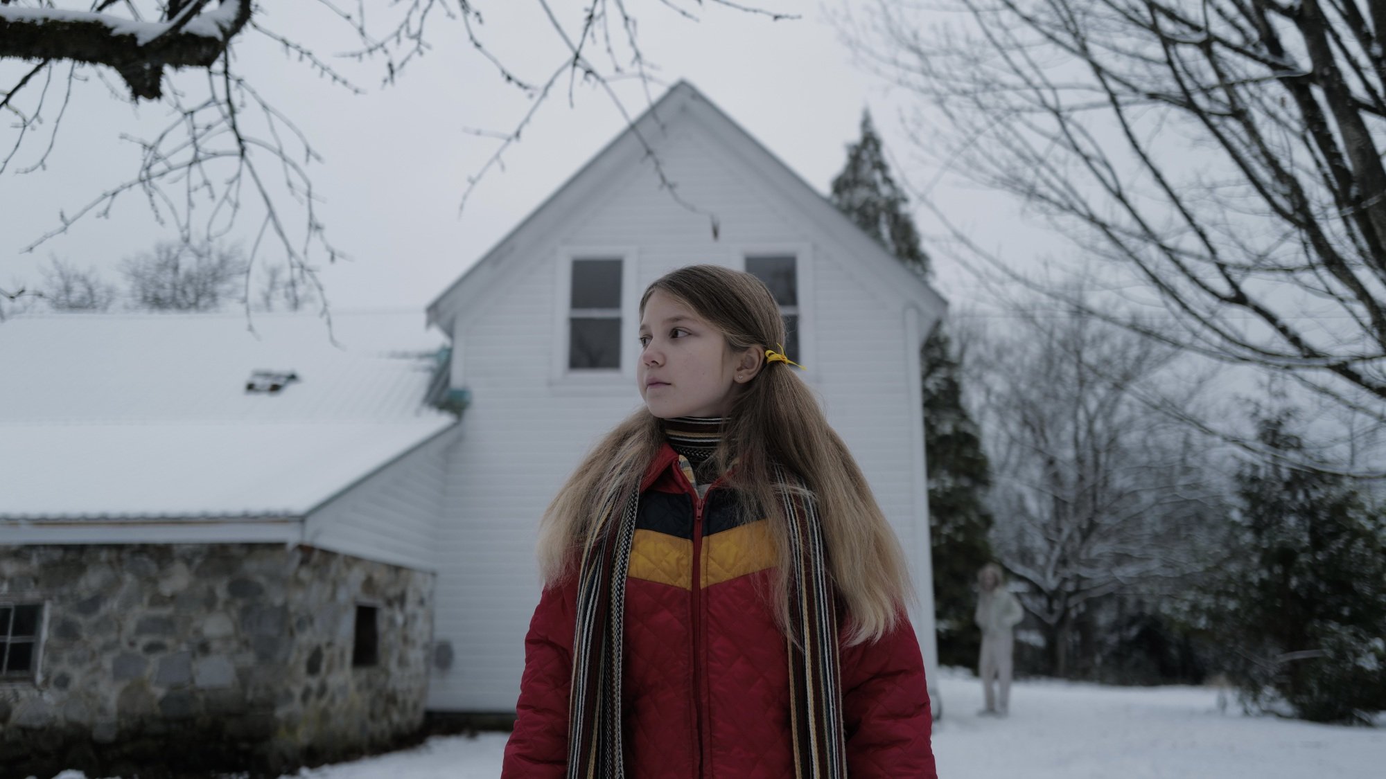 Une jeune fille vêtue d'un manteau d'hiver coloré se tient devant sa maison blanche dans la neige, tandis qu'un homme se cache au fond de l'arrière-plan.