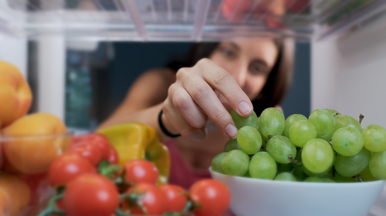 Femme atteignant un réfrigérateur rempli de produits frais, notamment des raisins et des tomates.