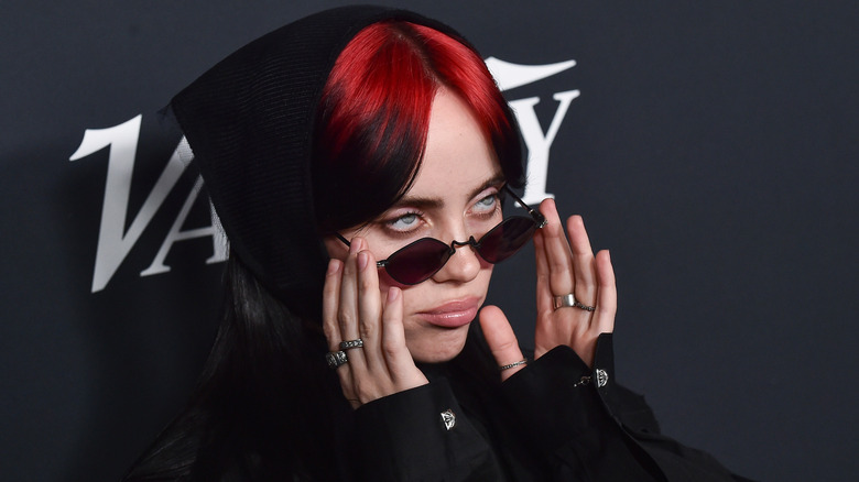 Billie Eilish avec des racines rouges et des lunettes de soleil, faisant la moue sur le tapis rouge