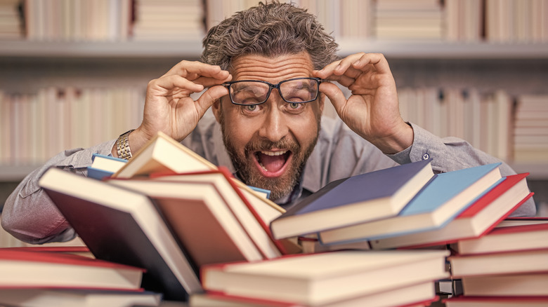 Un enseignant se tient derrière une pile de livres dans une bibliothèque en soulevant ses lunettes de son visage