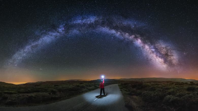 Une personne se tient sur une route avec une lampe de poche sous des millions d'étoiles et des nuages ​​de gaz violets de la Voie Lactée dans le ciel nocturne