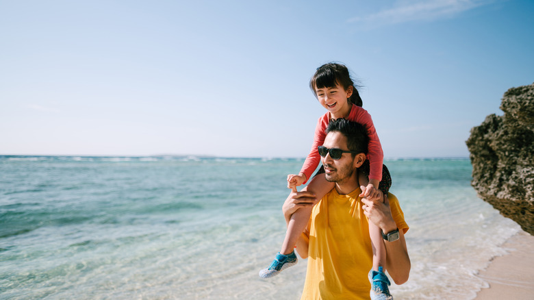 Un homme porte sa fille sur ses épaules sur une plage ensoleillée.