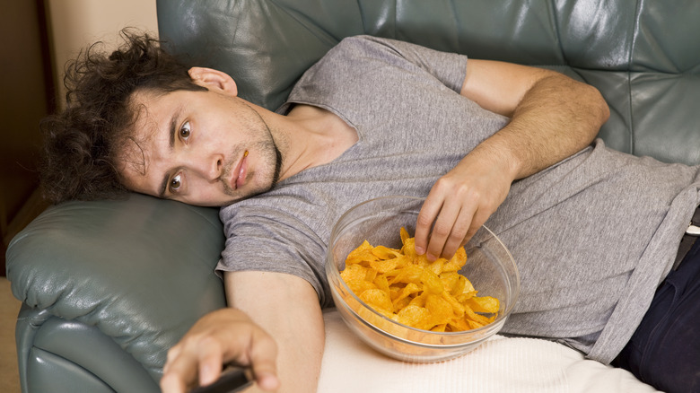 Un homme fatigué sur un canapé mangeant des croustilles.