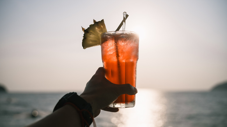 Une main tient un cocktail vers le soleil et l'eau