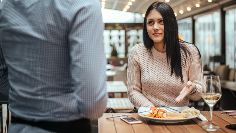 Une femme pointe vers son plat dans un restaurant tandis qu'une silhouette anonyme la regarde