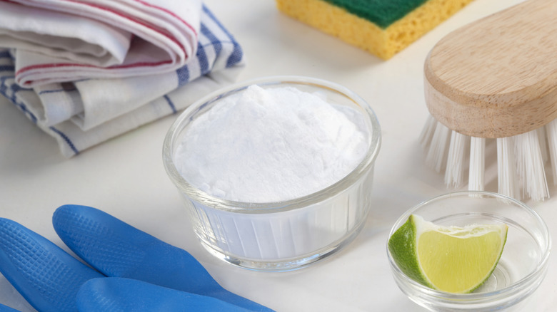 Petit bol de bicarbonate de soude entouré de gants, d'outils de nettoyage et d'un coin de citron vert