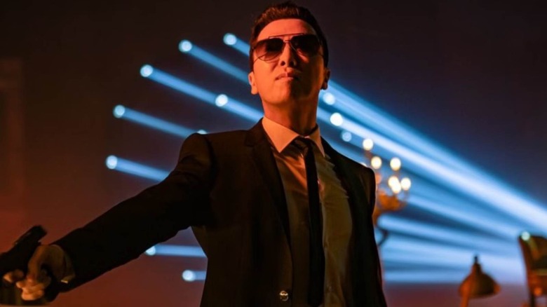 Caine de John Wick 4 portant des lunettes et un costume tenant un pistolet devant des trous de balles dans un mur