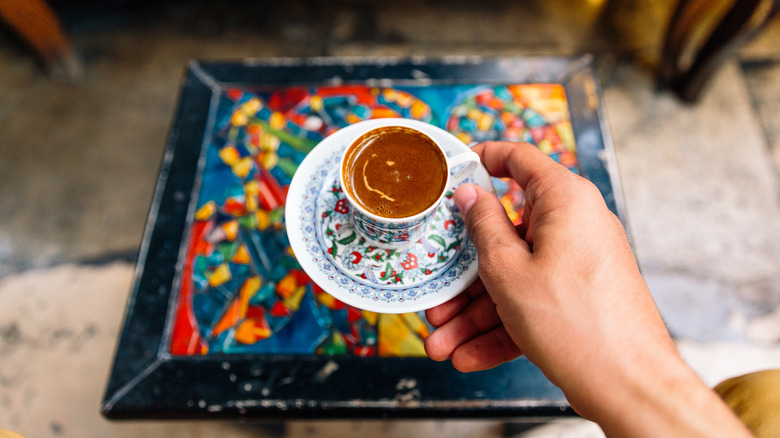 Une main tient une soucoupe et une tasse pleine de café turc sur une table colorée