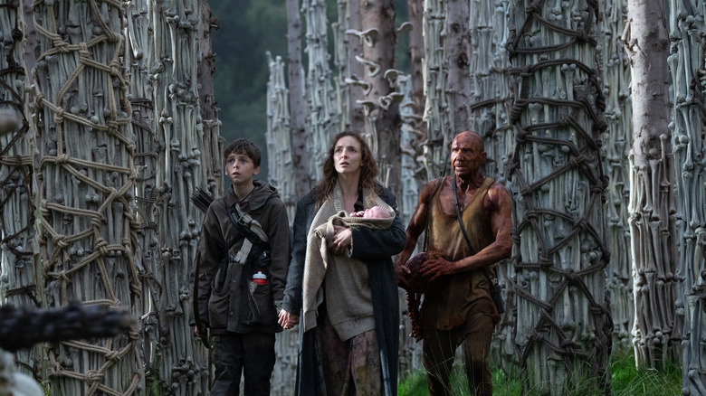 Spike, Isla (tenant un bébé) et le Dr Ian Kelson se tenant parmi de nombreux os dans 28 ans plus tard