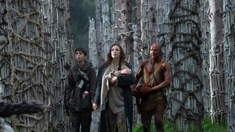 Spike, Isla (tenant un bébé) et le Dr Ian Kelson se tenant parmi de nombreux os dans 28 ans plus tard