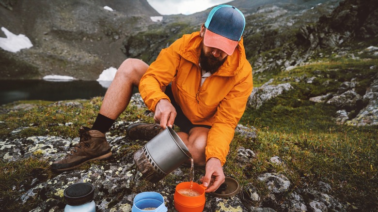 Un randonneur mâle verse de l'eau chaude dans une tasse d'orange à côté d'un lac alpin