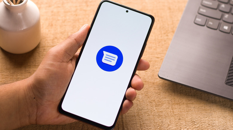 Une personne qui ait un téléphone avec le logo Google Messages en affichage; Un ordinateur portable et un petit vase blanc sont visibles à l'arrière.