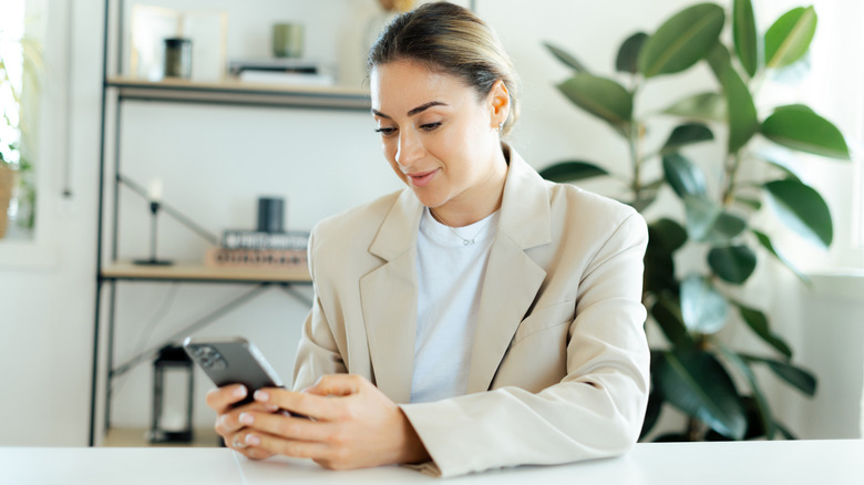 Une femme en tenue d'entreprise utilisant son téléphone alors qu'elle était assise sur un bureau avec des plantes et une étagère de différents articles est vue à l'arrière.