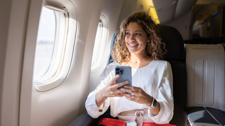 Une femme latine aux cheveux bouclés tenant son téléphone souriant et regardant par fenêtre d'avion