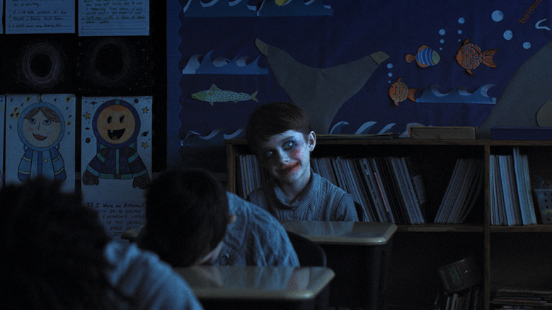 Alex Lilly avec un maquillage effrayant se trouve dans une salle de classe sombre tandis que d'autres élèves ont la tête sur leur bureau dans les armes