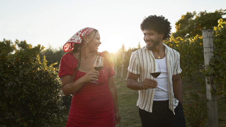 Couple souriant avec du vin rouge marche au milieu des plantes à la ferme pendant le coucher du soleil