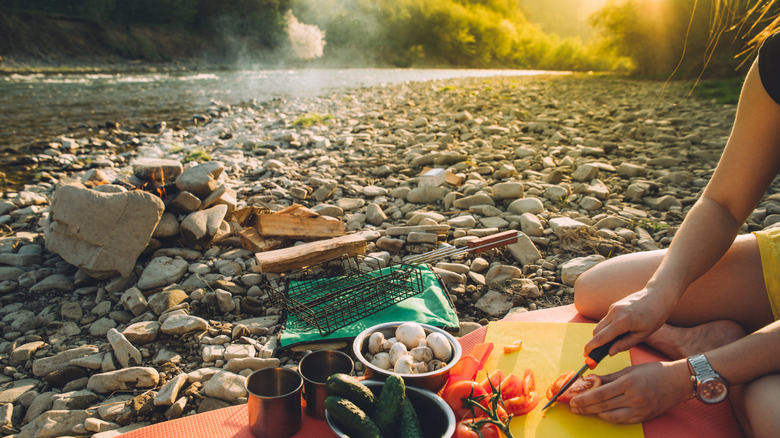 Les mains d'une femme sont montrées en train de couper des légumes tout en étant assis le long d'un lit de rivière rocheux