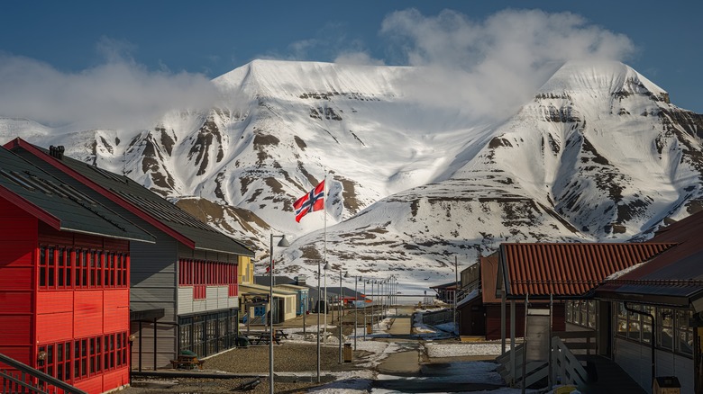 Une petite ville avec des bâtiments en bois colorés, un drapeau norvégien surélevé et des montagnes couvertes de neige en arrière-plan