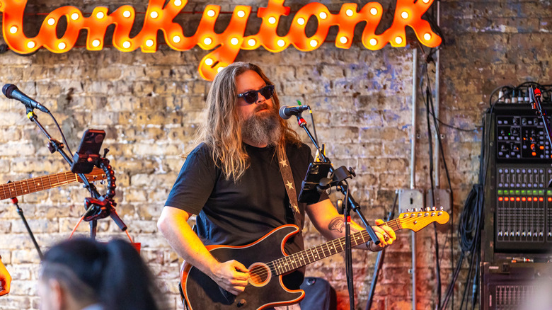 Un homme barbu aux cheveux longs jouant une guitare tout en se tenant sur scène avec des lunettes de soleil