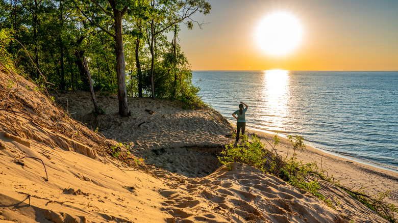 Une femme se tient sur des dunes regardant le coucher du soleil sur le grand lac, le sable en pente et les arbres sur le rivage