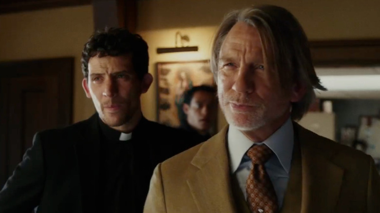 Benoit en costume marron souriant avec Jud et Cy derrière lui dans Wake Up Dead Man