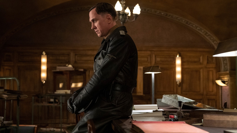 Lars Eidinger dans le rôle de Reinhold von Rumpel, assis contemplativement sur un bureau, dans All the Light We Cannot See