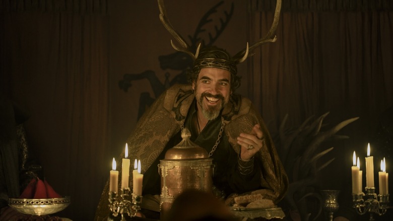 Daniel Ings dans le rôle de Lyonel Baratheon portant une couronne de bois de cerf et assis à une table dans Le Chevalier des Sept Royaumes