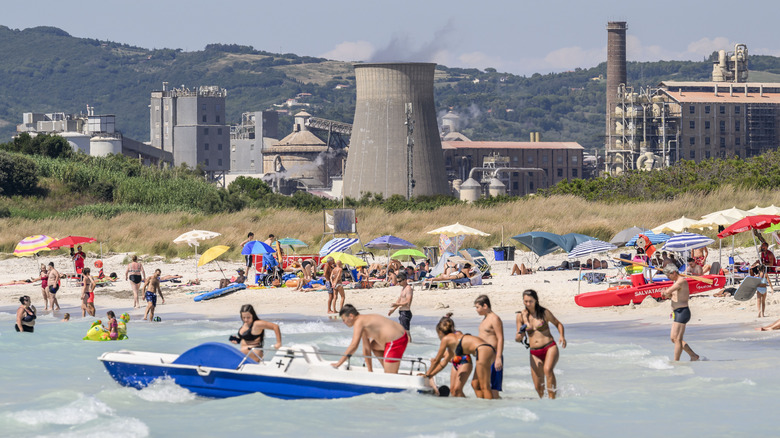 Gens jouant dans les eaux peu profondes d’une plage avec l’usine chimique en arrière-plan